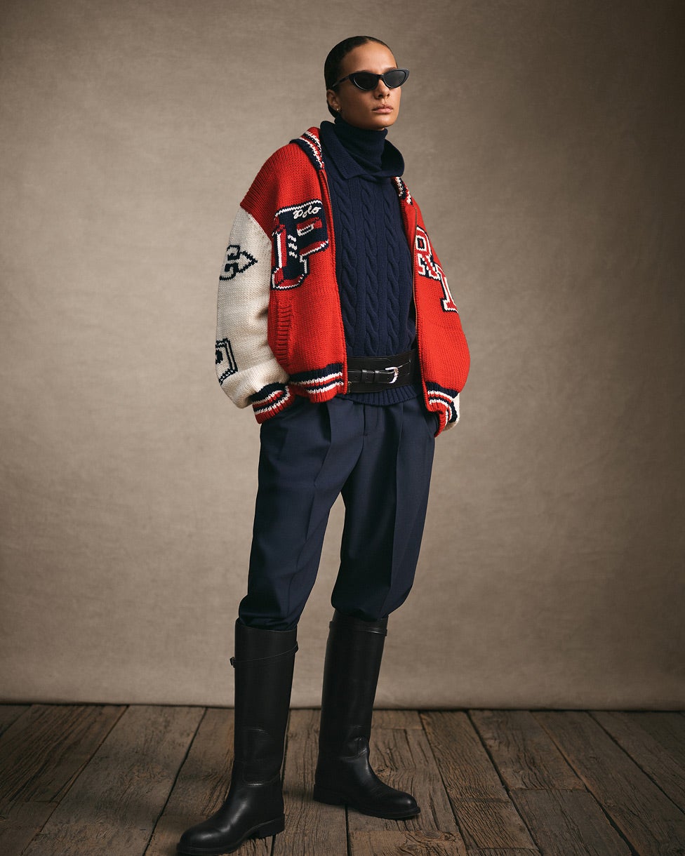 Femme debout portant une veste universitaire rouge et blanche, un pull et un pantalon marine et de hautes bottes noires.